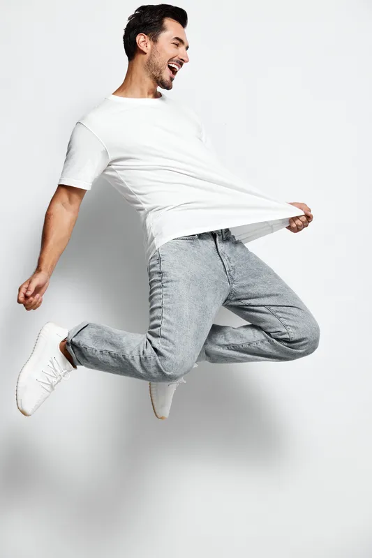 Happy energetic young man jumps joyfully in a white t-shirt and gray jeans, expressing excitement and positivity against a light solid background. People lifestyle concept.