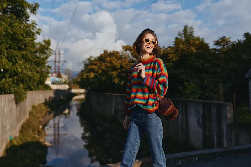 Young woman in her early 20s wearing a colorful striped sweater and sunglasses, smiling while walking by an urban canal in jeans with a brown leather bag, sunny lifestyle travel scene.