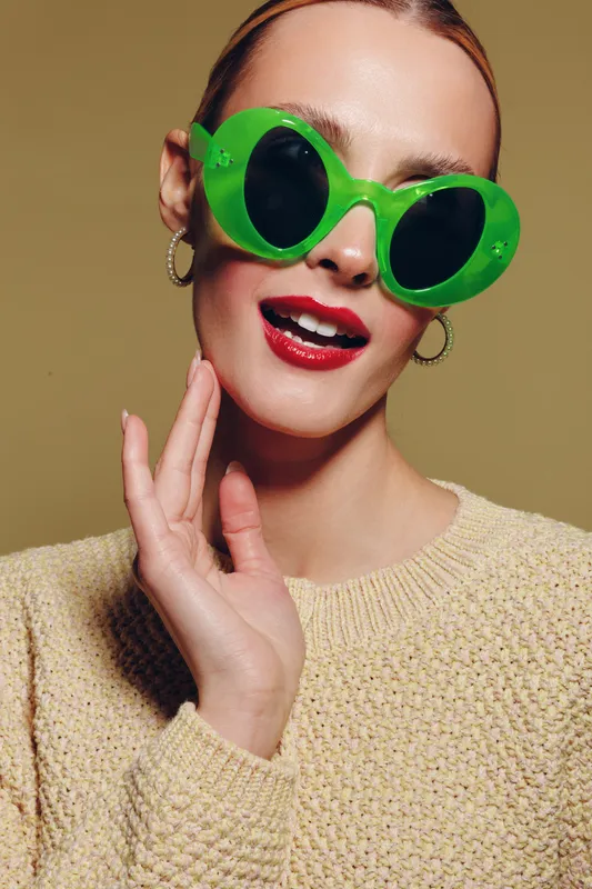 Young woman wearing large green sunglasses and red lipstick poses with hand near face against beige background. Fashion portrait with trendy accessories and stylish makeup.