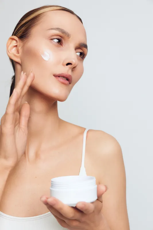 Young woman with blonde hair applies moisturizing cream to her cheek while holding a white jar in a bright studio setting, showcasing skincare and beauty routine.