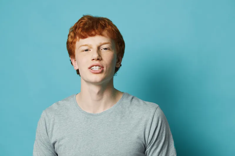 Young man with red hair and light skin posing in gray t shirt on blue background. Male portrait with casual clothing and confident facial expression in studio.