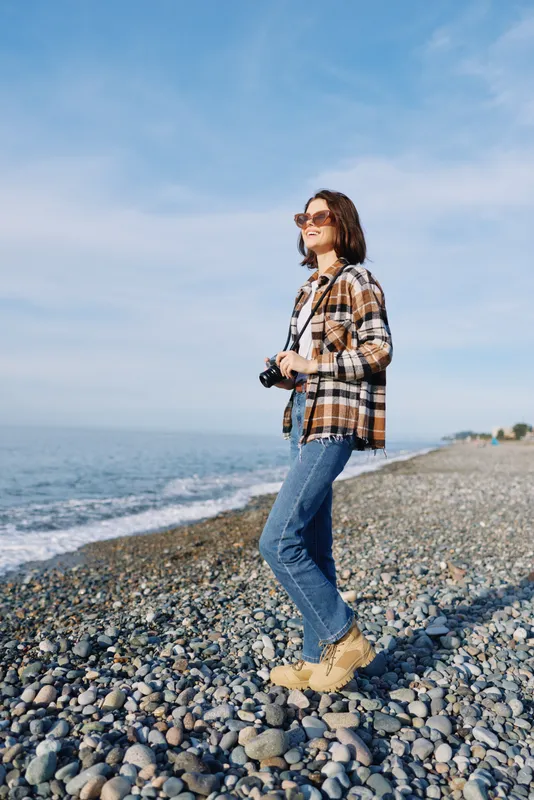 Casual photographer on a rocky beach with a camera, wearing a plaid shirt, jeans and sunglasses, enjoying the seaside landscape and clear sky.