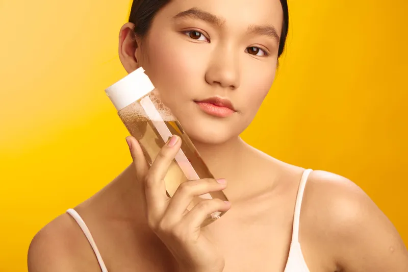 Asian woman holding a skincare bottle in a bright studio, showcasing a healthy glow. She smiles softly, presenting a calm, modern beauty moment and confident self care routine.