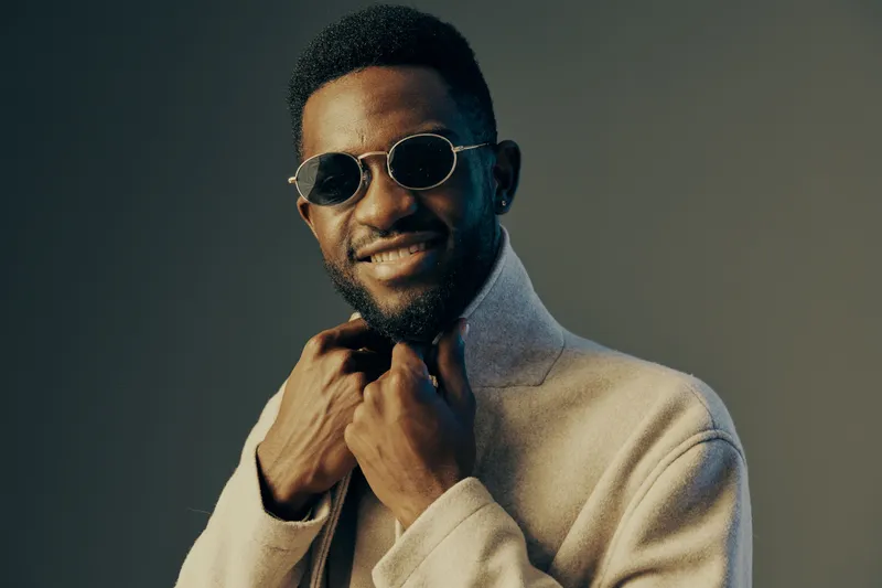 Stylish young African man in sunglasses smiles confidently, wearing a fashionable coat against a neutral background, showcasing contemporary fashion and joy