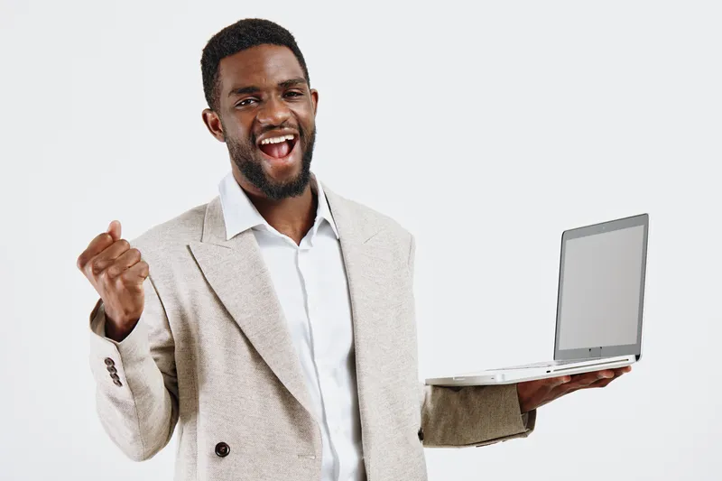 Confident smiling man of African descent holding a laptop computer in one hand, pointing upwards with the other, dressed in elegant beige blazer and white shirt, against plain white background.