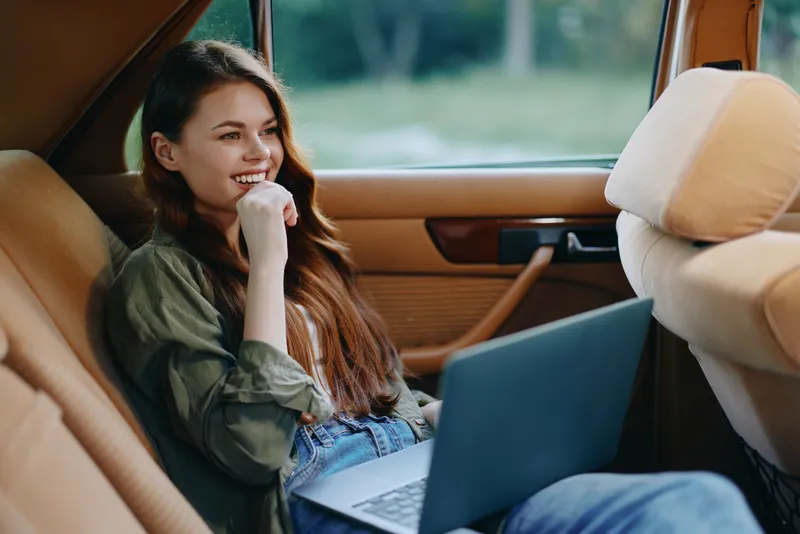 Young woman using a laptop in a car, smiling with a relaxed expression, showcasing remote working lifestyle in a natural setting.