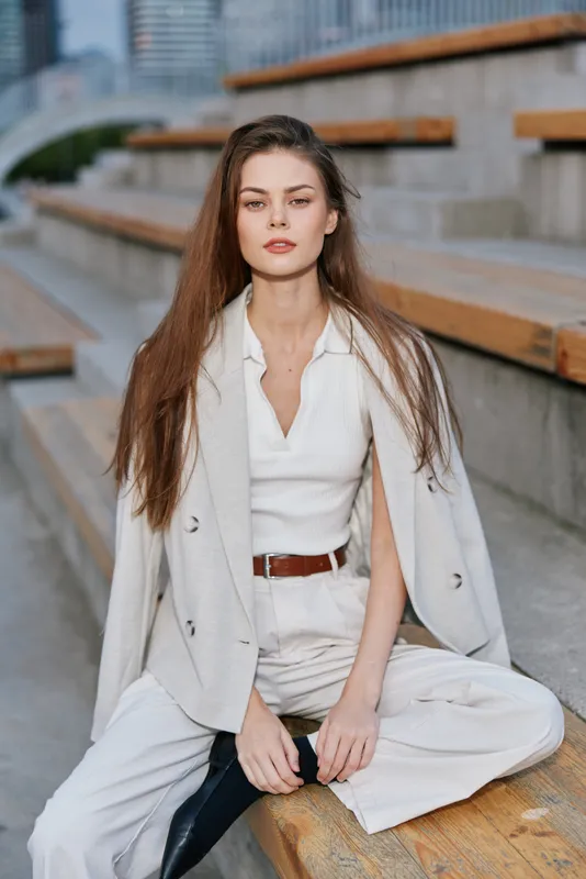 Young woman with long brown hair wearing beige business casual outfit sitting on outdoor wooden stairs with confident expression in urban setting, fashion style