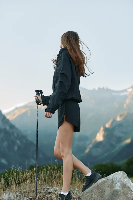 A solitary hiker stands on rocky terrain with a trekking pole, gazing at towering mountains. She wears a dark jacket and shorts, embodying adventure and freedom in a dramatic alpine landscape.