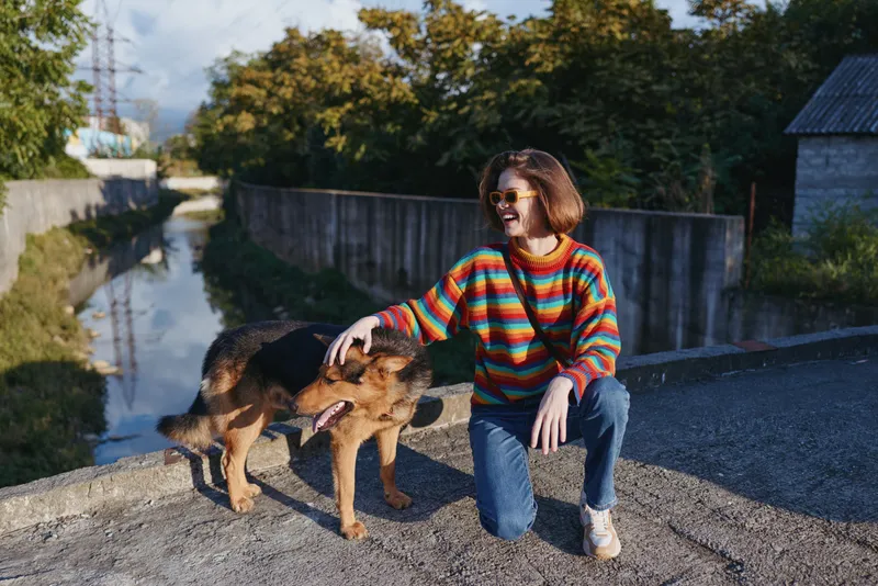 Woman in her early 20s wearing a colorful striped sweater and casual outfit, smiling while petting a German shepherd by a suburban canal on an autumn afternoon.