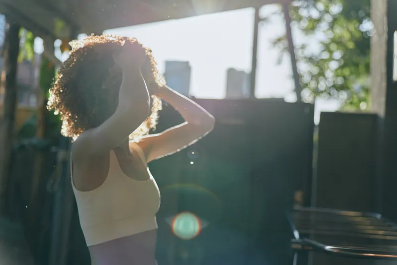 Young woman with curly hair adjusting in sunlight, wearing white tank top outdoors, natural light, green trees, casual lifestyle in warm atmosphere.