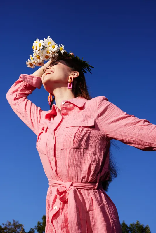 A joyful woman in a pink shirt dress with floral headwear poses against a bright blue sky, capturing a feeling of summer and nature, ideal for seasonal themes