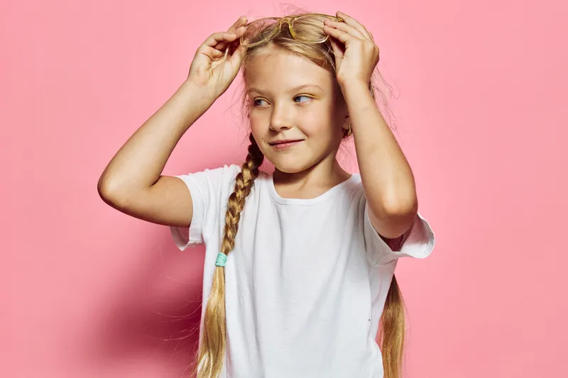 Blond girl with braids smiles gently adjusting glasses on pink background, wearing white t-shirt. Soft lighting studio portrait.