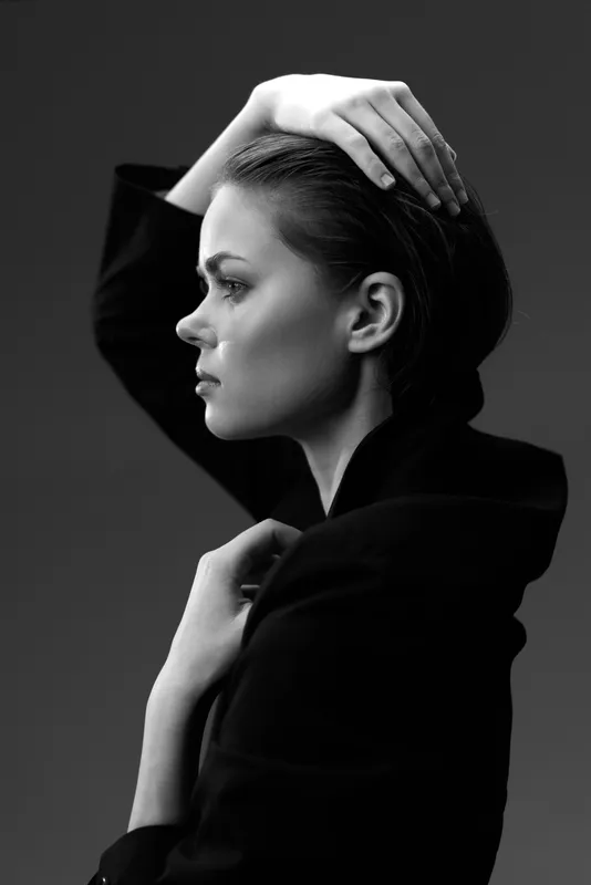 Black and white portrait of a thoughtful woman with hand on head wearing dark clothing on dark background