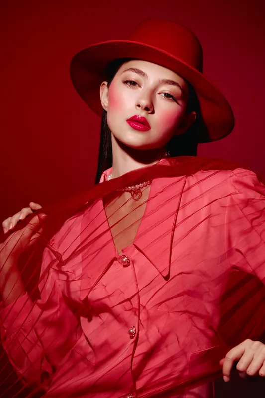 Fashionable woman wearing a stylish red hat and textured pink blouse poses confidently against a vibrant red background. Concept of beauty, fashion, creativity, and elegance.