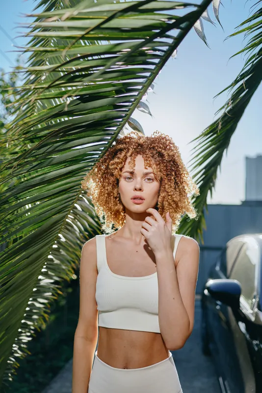 curly woman with natural hair in casual white outfit stands near palm leaves under bright daylight, showing relaxed summer style and fresh outdoor vibe.