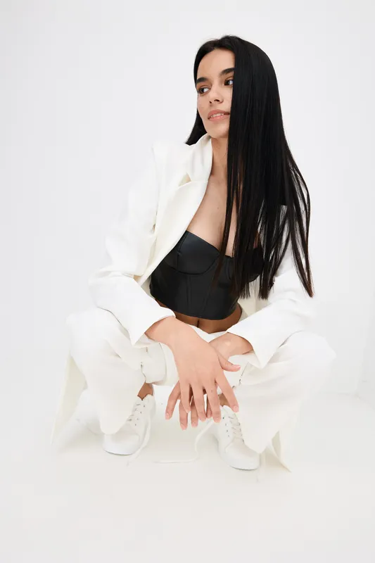 young woman crouching in stylish white suit and black top with long straight black hair, minimalistic bright background, modern fashion portrait composition
