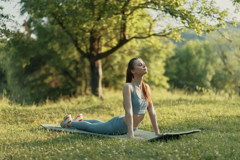 Woman practicing yoga in nature, demonstrating a cobra pose on a yoga mat in a serene outdoor setting surrounded by greenery and sunlight
