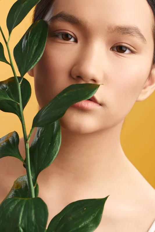 asian woman gazes softly at the camera, leaves framing her face in a studio portrait, warm yellow backdrop creating a calm, intimate and artistic mood.