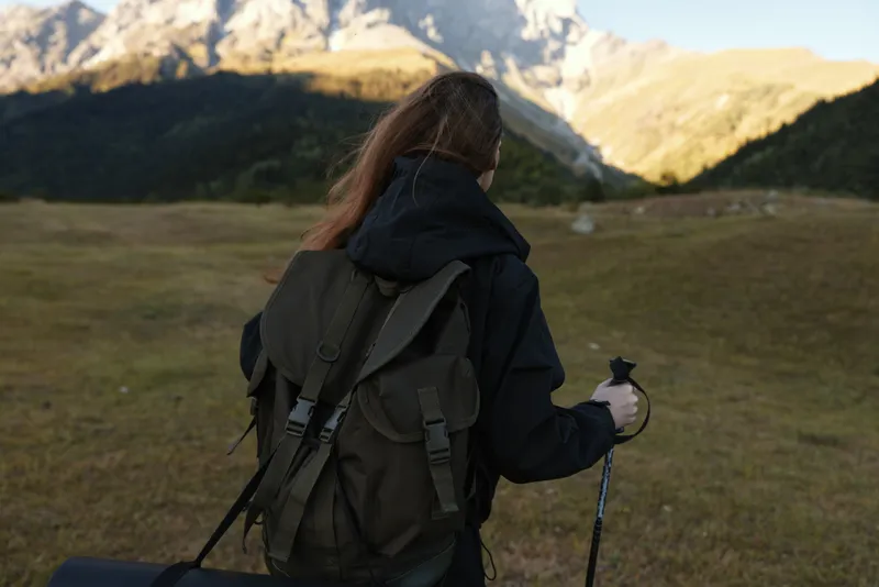 A lone hiker with a sturdy backpack walks through a sunlit meadow toward rugged mountains, conveying adventure, preparation, and outdoor exploration in a serene landscape.