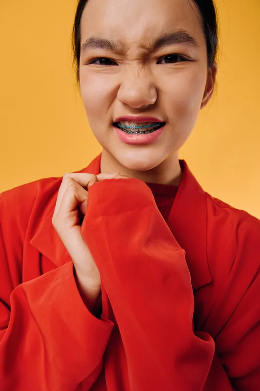 Young woman with braces is making a grimace face wearing a red jacket on orange background. Closeup portrait showing expression, fashion and teeth with metal braces visible.
