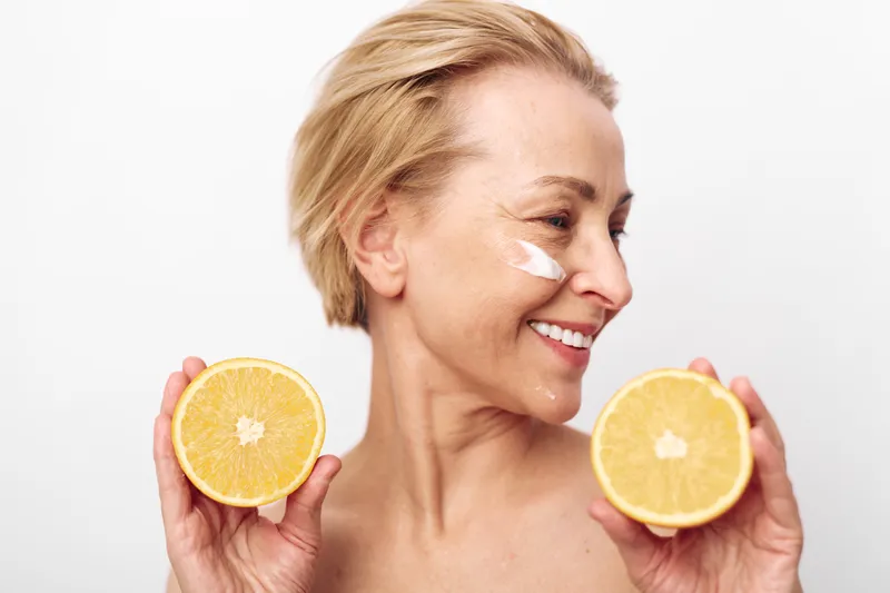 Smiling mature woman holds two halves of grapefruit near face with cream on cheek, isolated on white background. Concept for skincare, natural beauty, healthy lifestyle, and wellness.