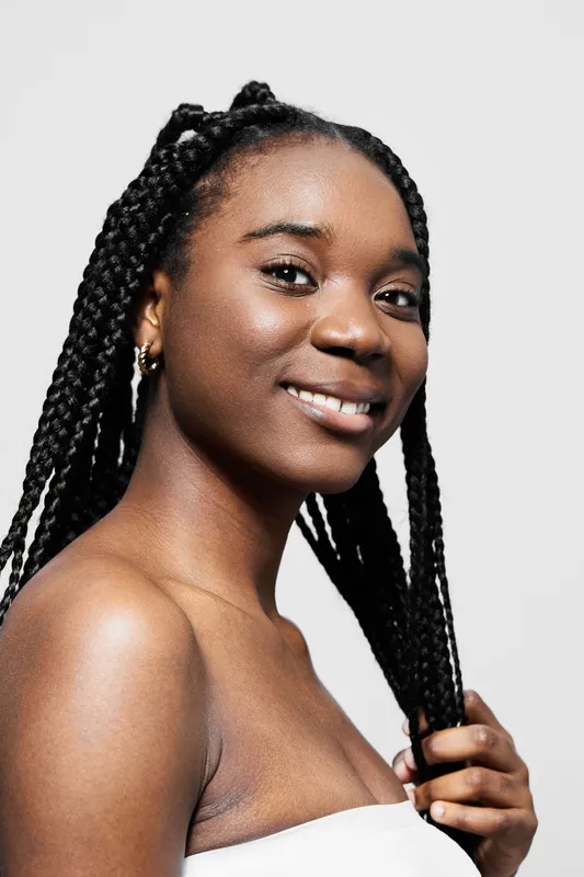 Smiling African woman with braided hair wearing a white top, showcasing natural beauty and confidence against a neutral background for beauty and skincare concepts