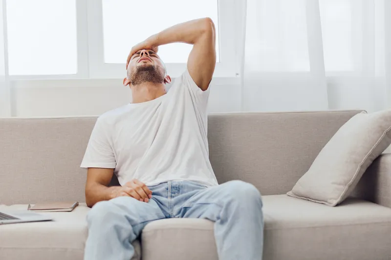 Depressed man sitting on a couch, reflecting on life challenges, dressed in casual clothing, expressive emotional moment captured in bright natural light.