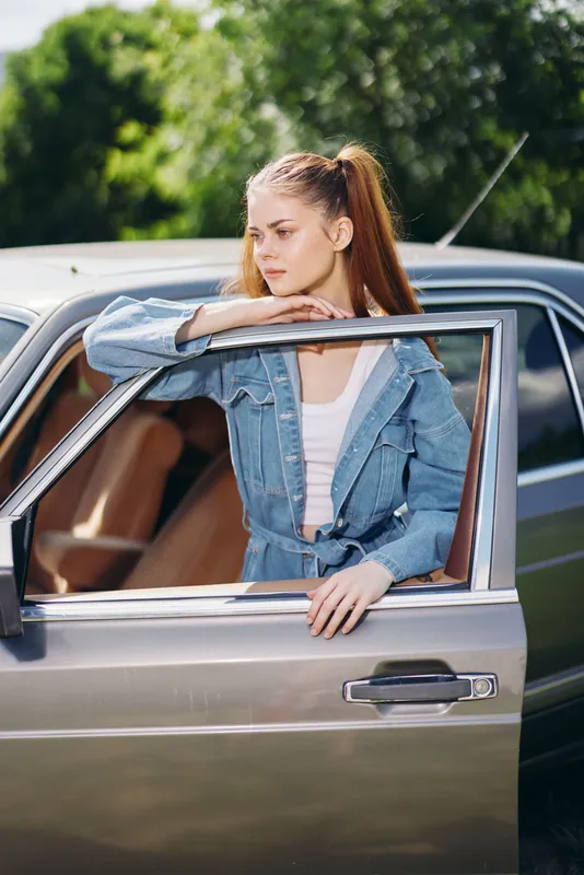 Young woman in denim outfit leaning out of a car window, enjoying a sunny day outdoors with lush green background, conveying a sense of freedom and adventure.
