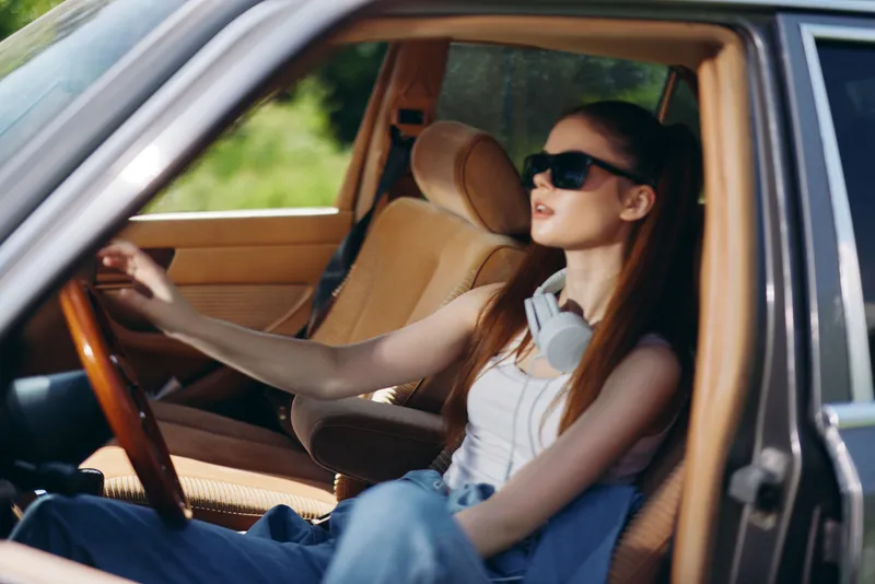 Young woman in casual attire wearing sunglasses and headphones, sitting in a vintage car, exuding confidence and style against a lush green backdrop.