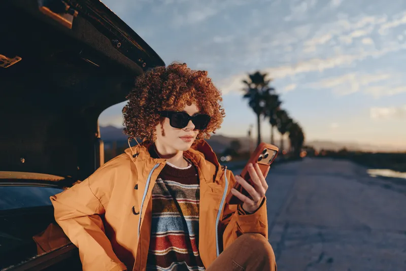 Woman lifestyle outdoors, curly hair, sunglasses, wearing orange jacket, sitting by car with open trunk, holding smartphone, relaxed pose, sunset sky, warm evening light, casual street style