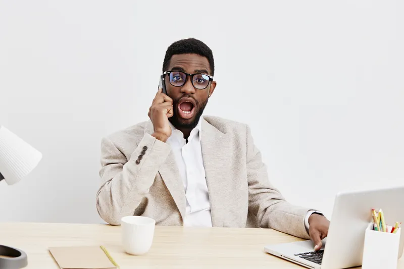 Confused young man with glasses talking on mobile phone while sitting at desk in modern office, surprised expression, casual blazer, white shirt, work environment, indoor shot, colorful background, bu