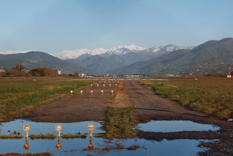Tranquil rural landscape with a muddy path crossing a wet field and reflective pools, distant snowcapped mountains under a clear blue sky, bright morning light.