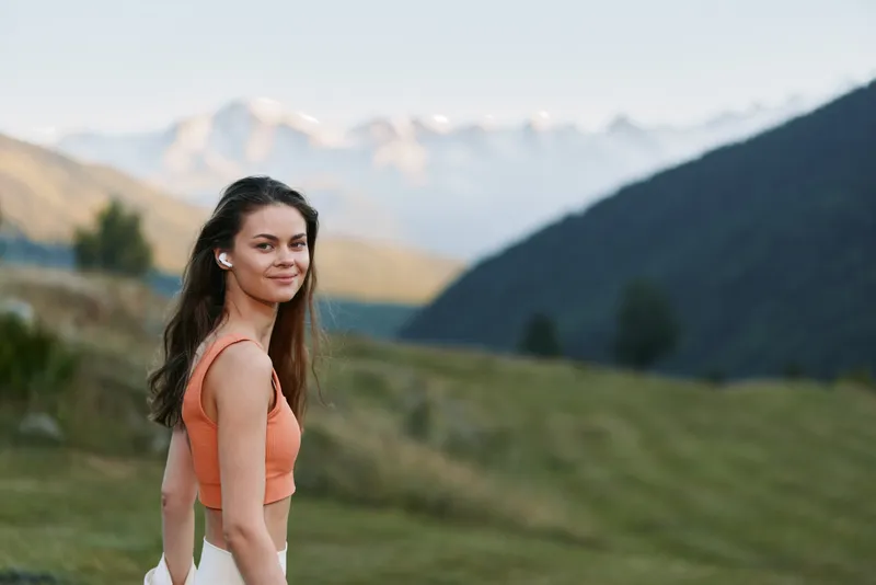 A cheerful woman stands outdoors in a scenic mountain landscape. She smiles warmly, wearing a coral top, exuding confidence and a relaxed, adventurous vibe.