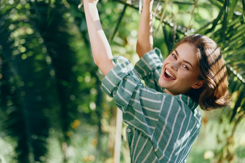 A joyful woman smiles brightly as she reaches up on a leafy branch in a vibrant outdoor scene, conveying energy, optimism, and carefree happiness amid nature.