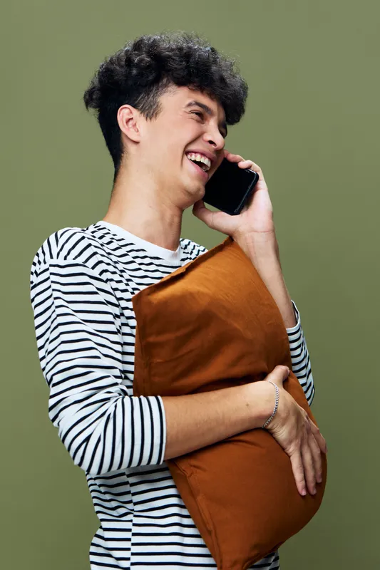Young man smiling while talking on smartphone, holding a brown pillow, wearing striped shirt against olive green background, joyful and casual atmosphere.