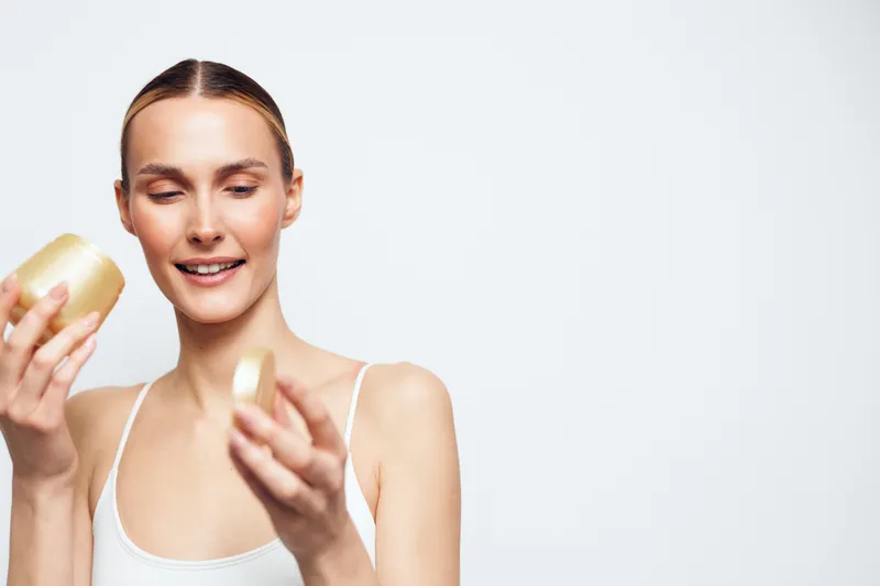 Young woman applying face cream for skincare routine, holding a small jar, smiling softly, focused on beauty and hygiene, spa style care, clean light background.
