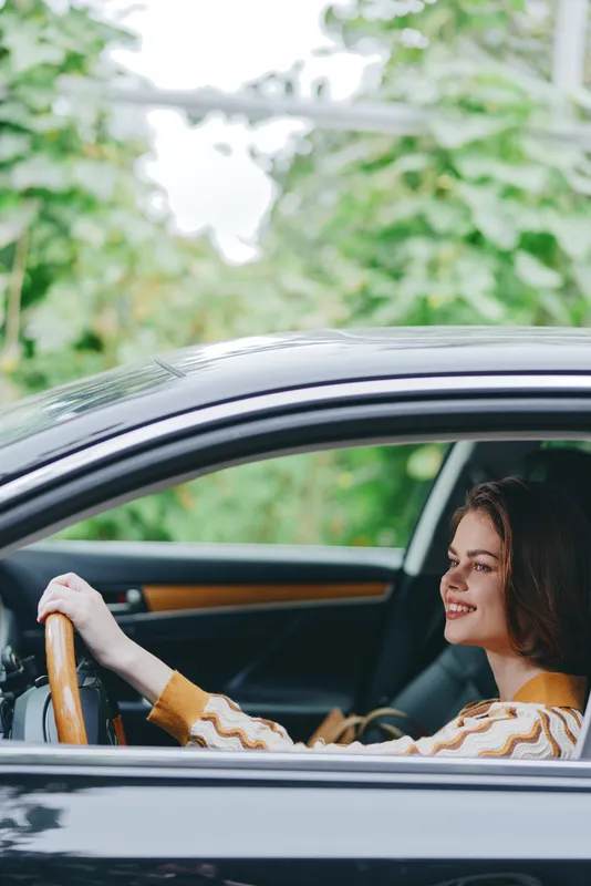 A woman sits behind the steering wheel of a sleek car, smiling softly as she drives along a calm street.