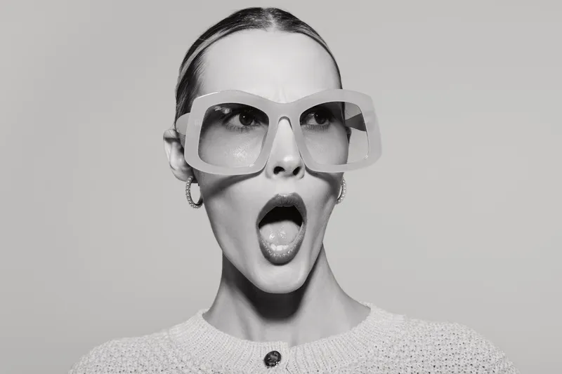 Surprised Caucasian woman with pale skin wears oversized sunglasses and light sweater, mouth open, posing against plain background. Concept of astonishment and fashion accessories.