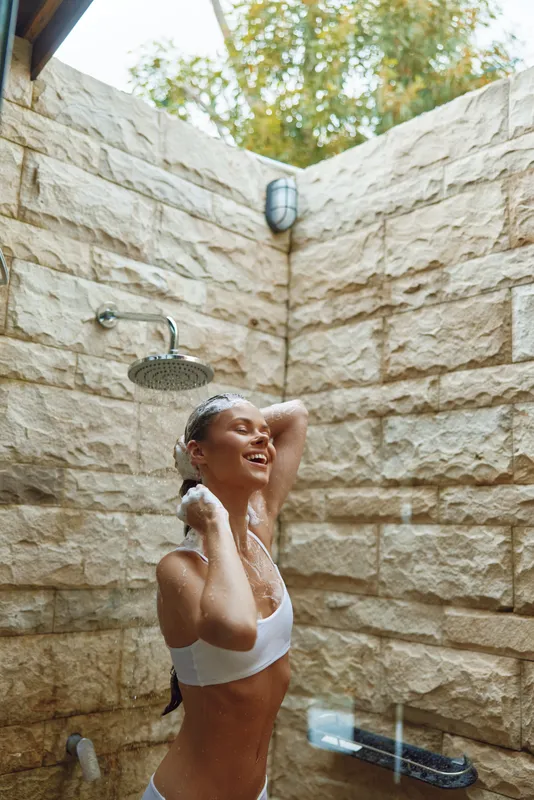 young woman enjoying outdoor shower with natural stone wall and greenery background, smiling and relaxed in bright daylight, summer wellness concept