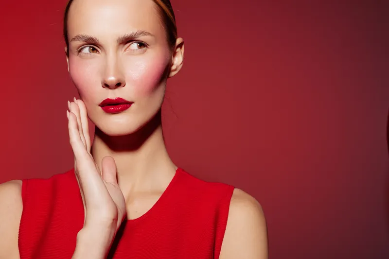 Portrait of a thoughtful woman with red lipstick and blush wearing a red sleeveless top against a red background. Concept of beauty, fashion, and makeup with a serious expression.