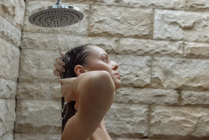 woman showering outdoors under rain showerhead with stone wall background, wet hair, natural morning light, relaxed mood in spa setting