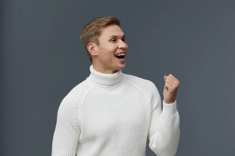 Joyful man smiling happily in casual white sweater with fist raised in celebration against plain gray background, expression of success and positive energy, studio portrait, people lifestyle concept.