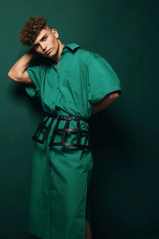Young man in fashion styled green dress with modern belt posing, curly hair and hand behind head on dark background for portrait and style concept photography.