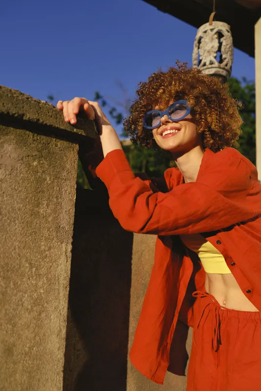 Smiling woman with curly hair in an orange jacket leaning on a wall outdoors. Casual lifestyle scene, sunny mood, warm colors, carefree moment, natural light