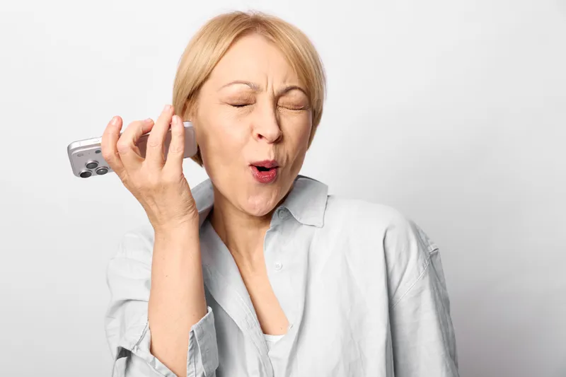 Stressed elderly woman with phone to ear, eyes closed, tense expression. Headache and anxiety appear during a call, highlighting health concerns today in a clean indoor portrait.