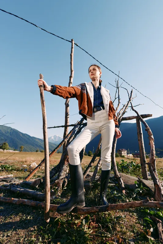 Woman standing in mountains wearing boots and jacket, holding a stick in a rural landscape. Outdoor portrait with confident pose, sunlight and scenic field backdrop.