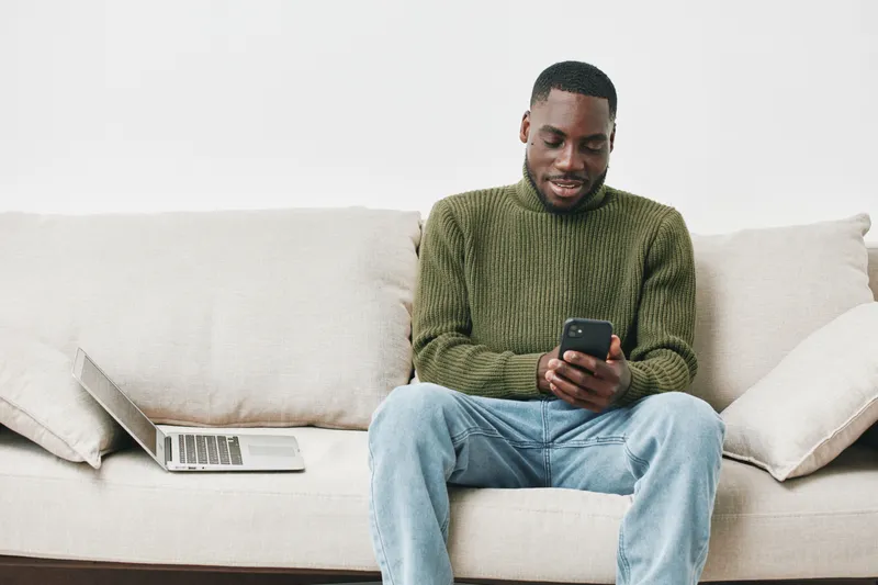 Young Black man sitting on a couch, smiling and using a smartphone, wearing a green sweater and jeans, with a laptop nearby, in a modern living room