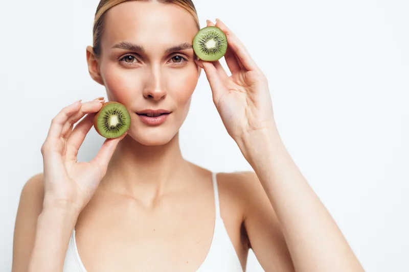 Bright studio portrait of a woman holding kiwi halves near her cheeks, fresh skincare moment, clear skin, soft light, natural tones, simple white background
