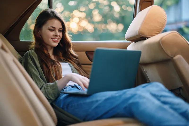 Young woman working on laptop in a car, smiling and relaxed, wearing casual clothing, with a soft green background, embodying a carefree and modern work-life balance.