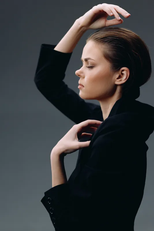 Profile of a thoughtful woman in black clothing with elegant pose and calm expression on dark background.